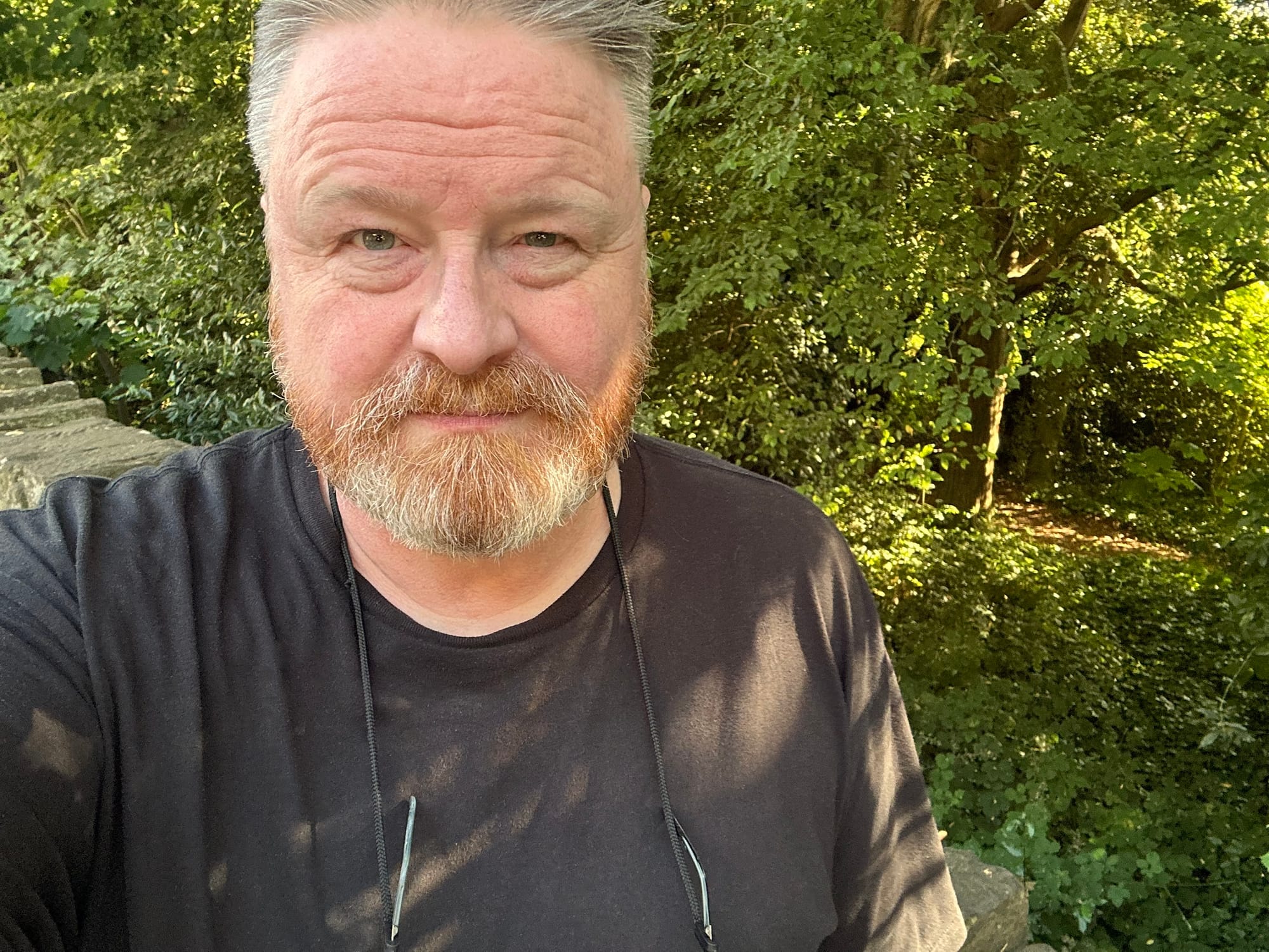 Gareth wearing a black t-shirt, standing in front of some trees, with the sunlight shining through their leaves.